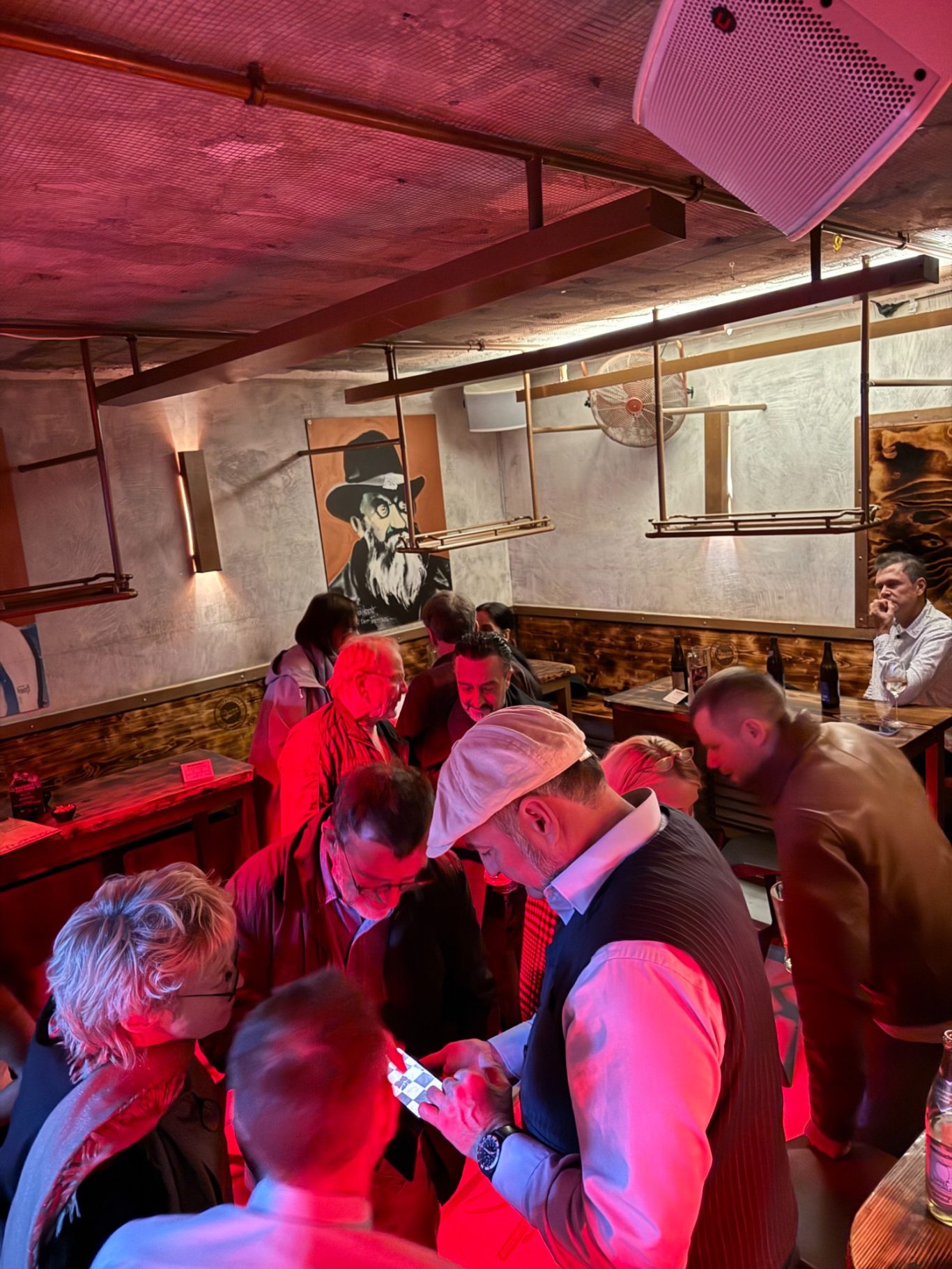 1. Unionsbäu-Stammtisch in der Boazn / 15.10.26 Mehrere Gäste stehen im rot beleuchteten Gastraum der Boazn zusammen und schauen gemeinsam auf ein Smartphone.