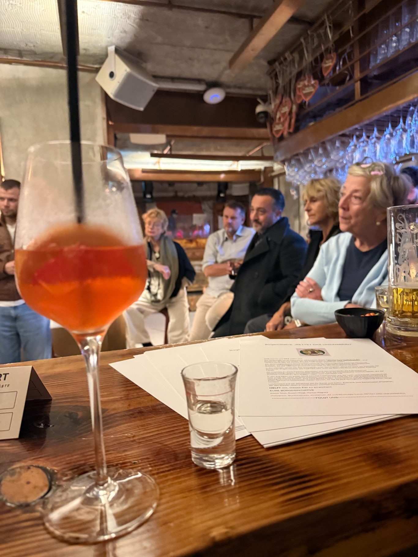 1. Unionsbäu-Stammtisch in der Boazn / 15.10.26 Im Vordergrund stehen ein Spritz-Glas und ein Schnapsglas auf der Theke, daneben liegt ein Stapel Infoblätter der Bürgerinitiative. Im Hinte