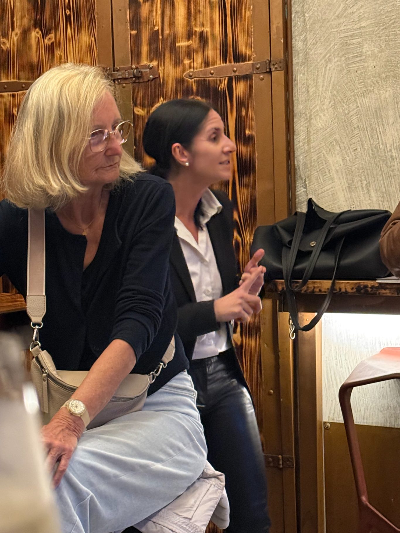 1. Unionsbäu-Stammtisch in der Boazn / 15.10.26 Zwei Frauen sitzen vor einer dunklen Holzwand; die eine hört aufmerksam zu, die andere erklärt etwas mit erhobener Hand.